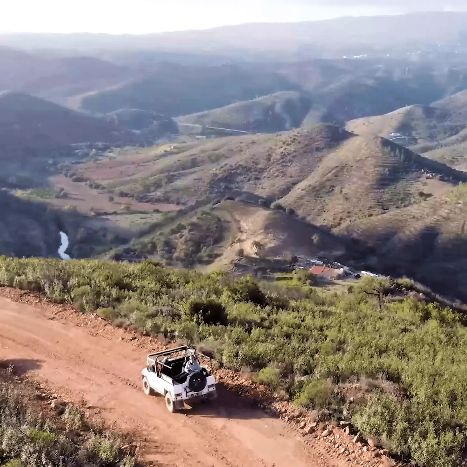 Drone view of the white jeep on a red earth track high above folded hills and a river valley